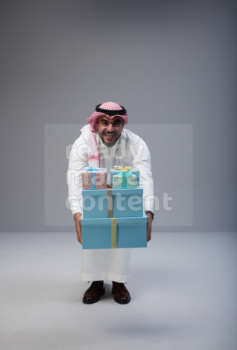 Saudi man Wearing Thob and red Shomag standing holding gift box on Gray background
