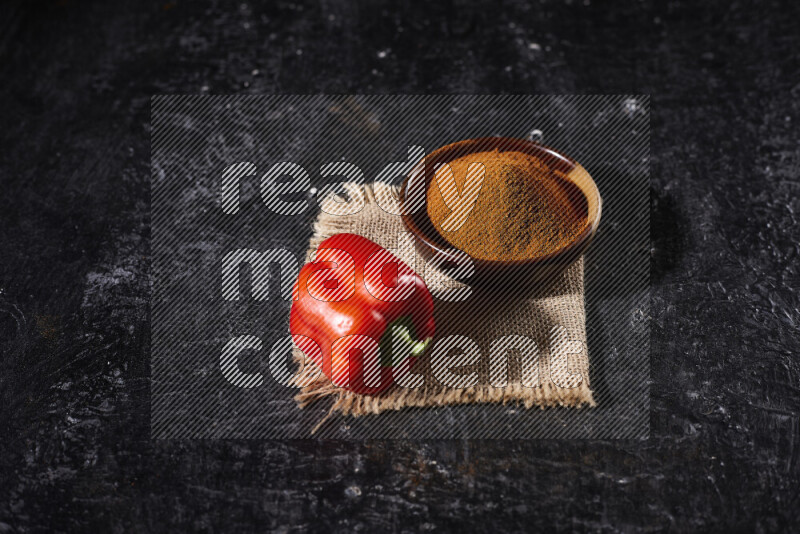 A wooden bowl full of ground paprika powder with a red bell pepper on a burlap fabric on black background