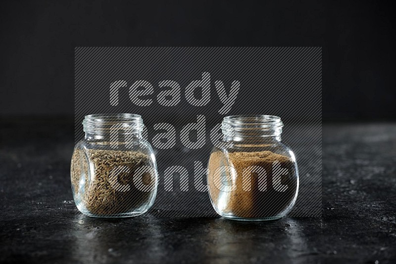 2 glass spice jars full of cumin powder and seeds on a textured black flooring