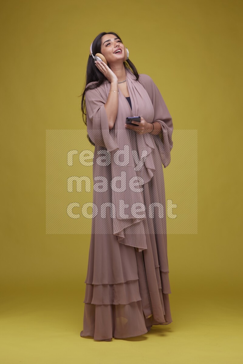 A Saudi woman Listing to Music on a Yellow Background wearing Brown Abaya