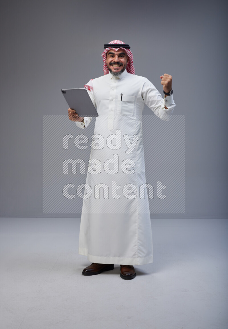 Saudi man Wearing Thob and red Shomag standing working on tablet on Gray background