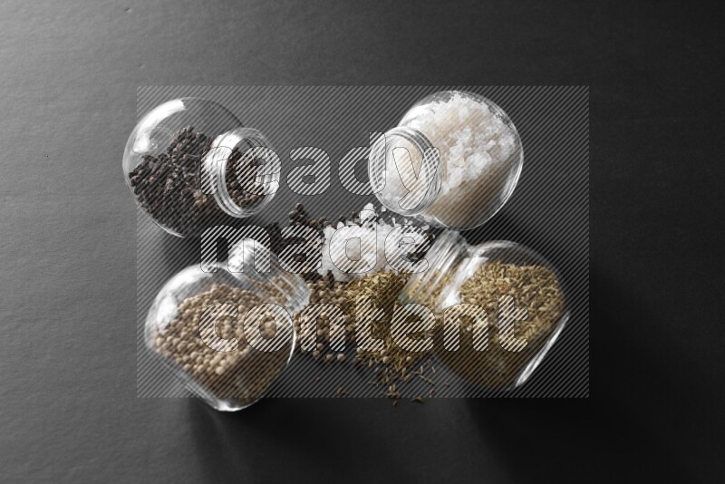 4 glass spice jars full of salt, black peppers, white peppers and cumin on black flooring