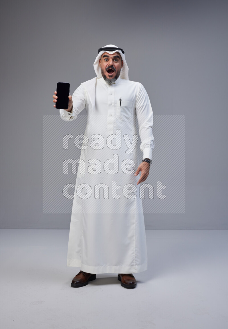 Saudi man Wearing Thob and white Shomag standing showing phone to camera on Gray background
