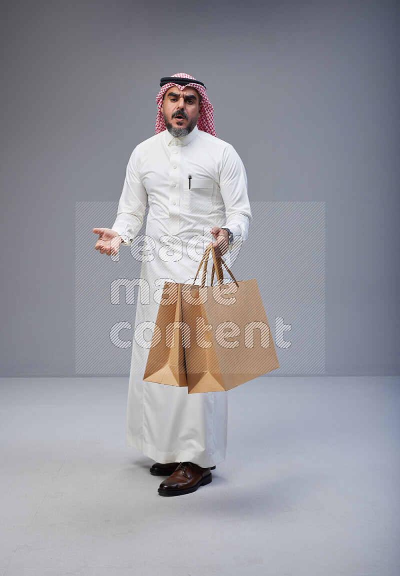 Saudi man Wearing Thob and red Shomag standing holding shopping bag on Gray background