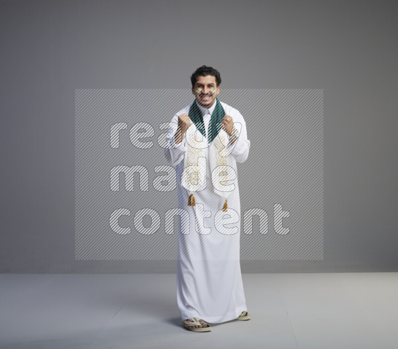 A Saudi man standing wearing thob and Saudi flag scarf with face painting on gray background