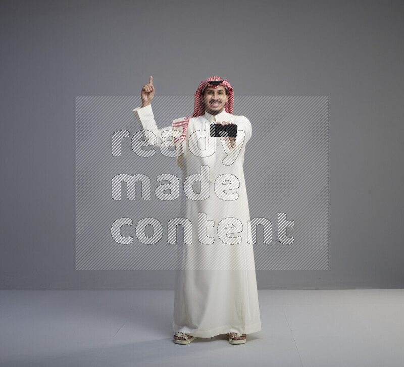 A Saudi man standing wearing thob and red shomag showing phone to camera on gray background