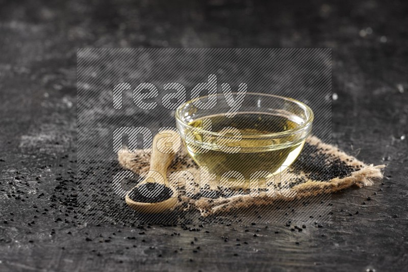 A glass bowl full of black seeds oil and wooden spoon full of black seeds with seeds spreaded on burlap fabric on a textured black flooring in different angles