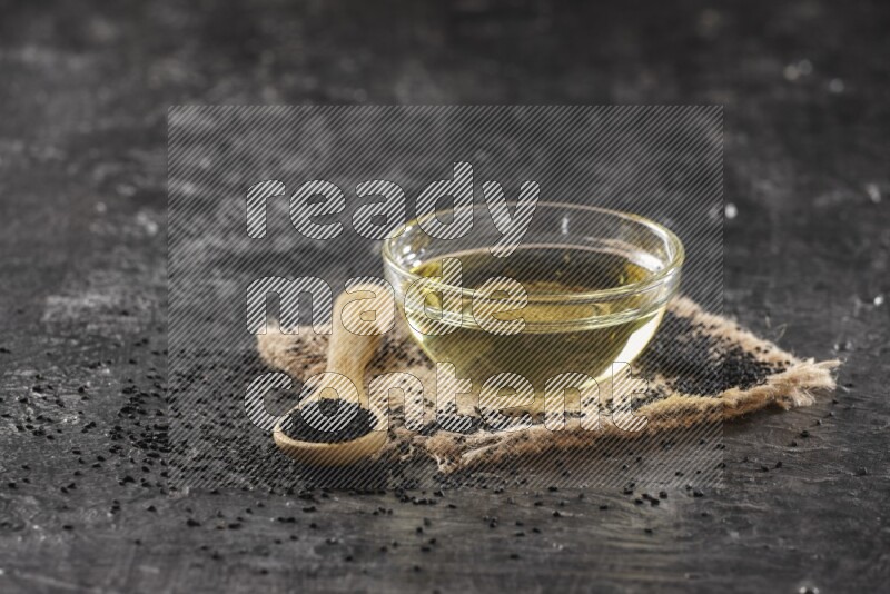 A glass bowl full of black seeds oil and wooden spoon full of black seeds with seeds spreaded on burlap fabric on a textured black flooring