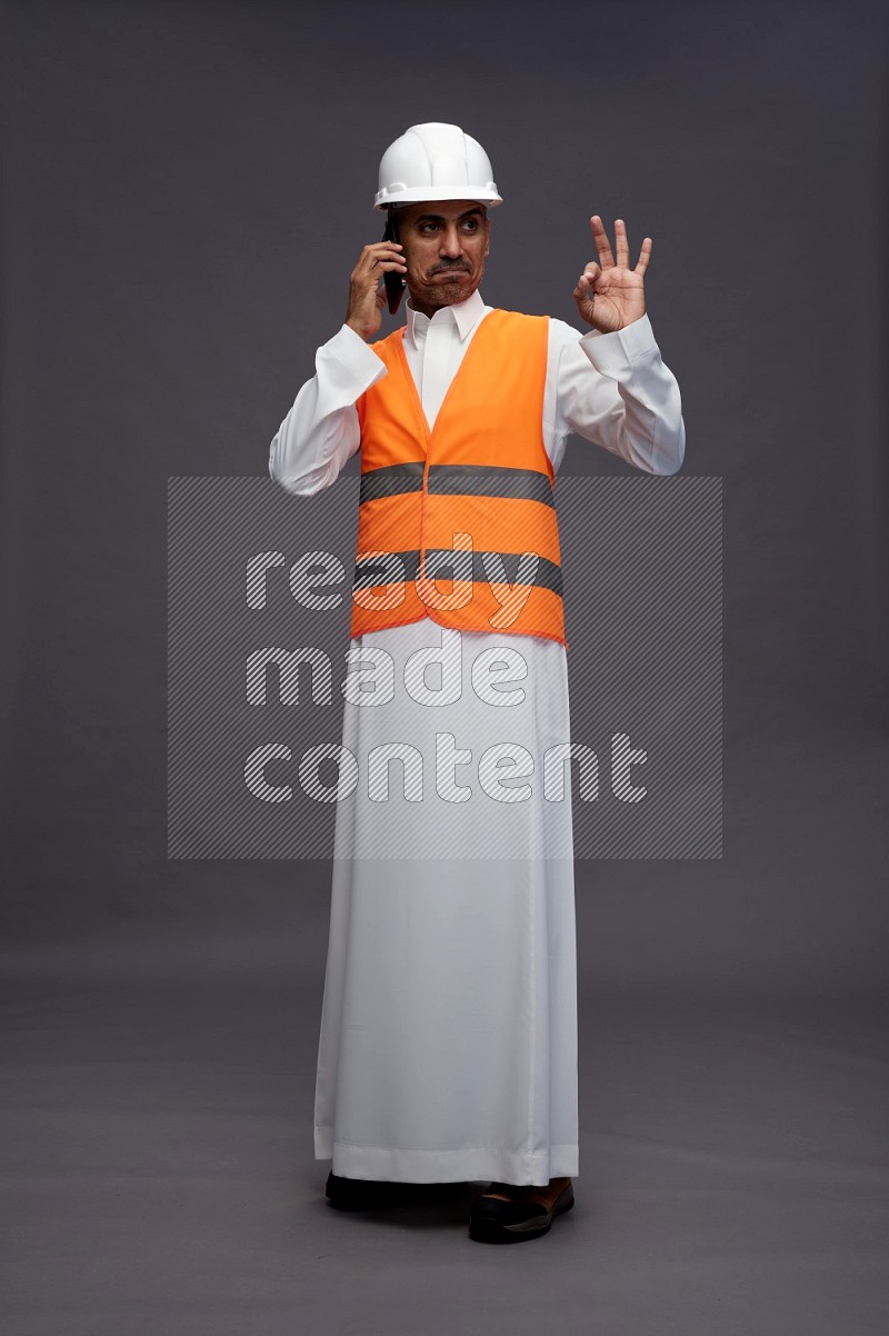 Saudi man wearing thob with engineer vest standing talking on phone on gray background