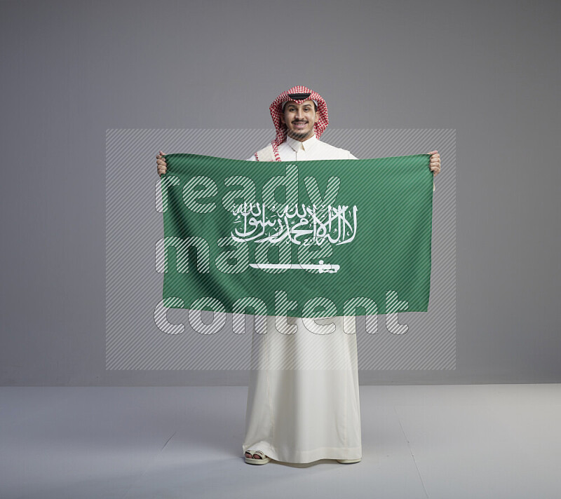 رجل سعودي يرتدي ثوب ابيض وشماغ احمر ويحمل علم كبير للسعودية