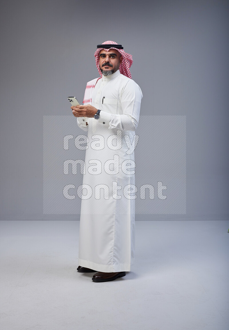 Saudi man Wearing Thob and red Shomag standing texting on phone on Gray background