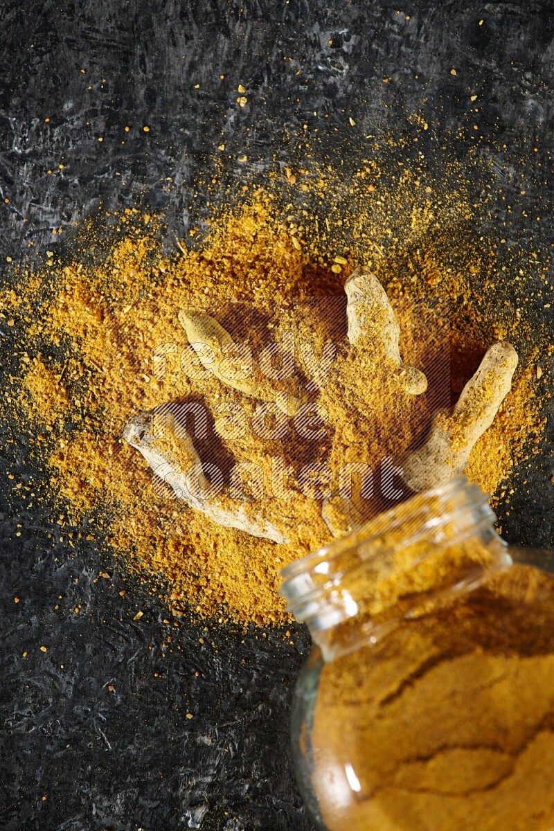 A flipped glass spice jar full of turmeric powder and powder spilled out of it with dried whole fingers on textured black flooring