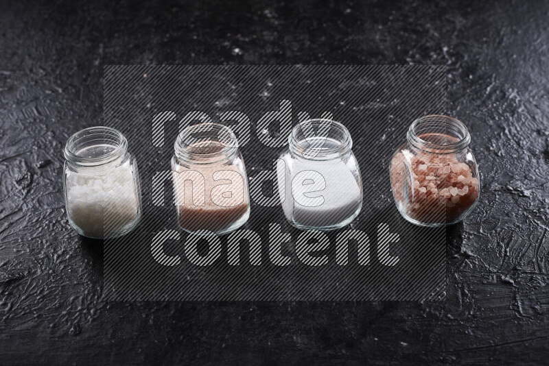 4 glass jars filled with table salt, coarse sea salt, fine himalayan salt and coarse himalayan salt on black background