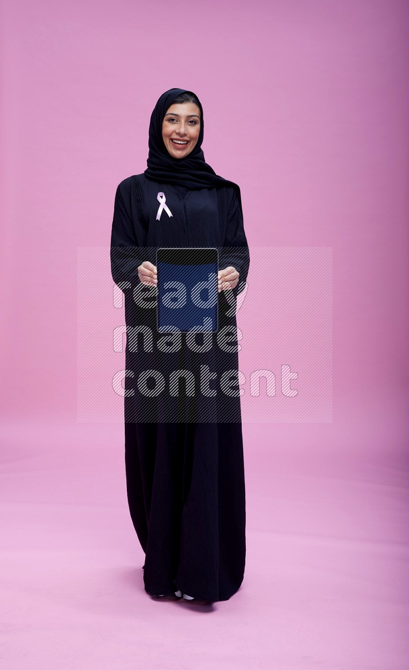 Saudi woman wearing pink ribbon on Abaya standing showing a tablet to camera on pink background