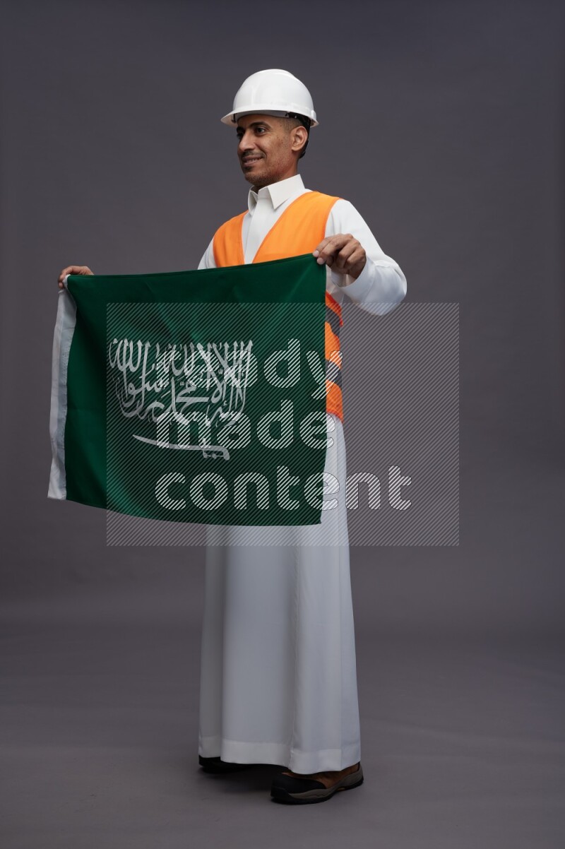 Saudi man wearing thob with engineer vest standing holding Saudi flag on gray background
