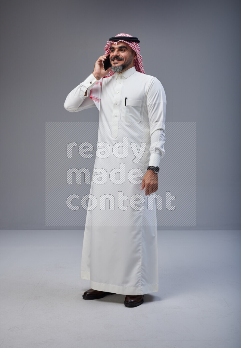 Saudi man Wearing Thob and red Shomag standing talking on phone on Gray background