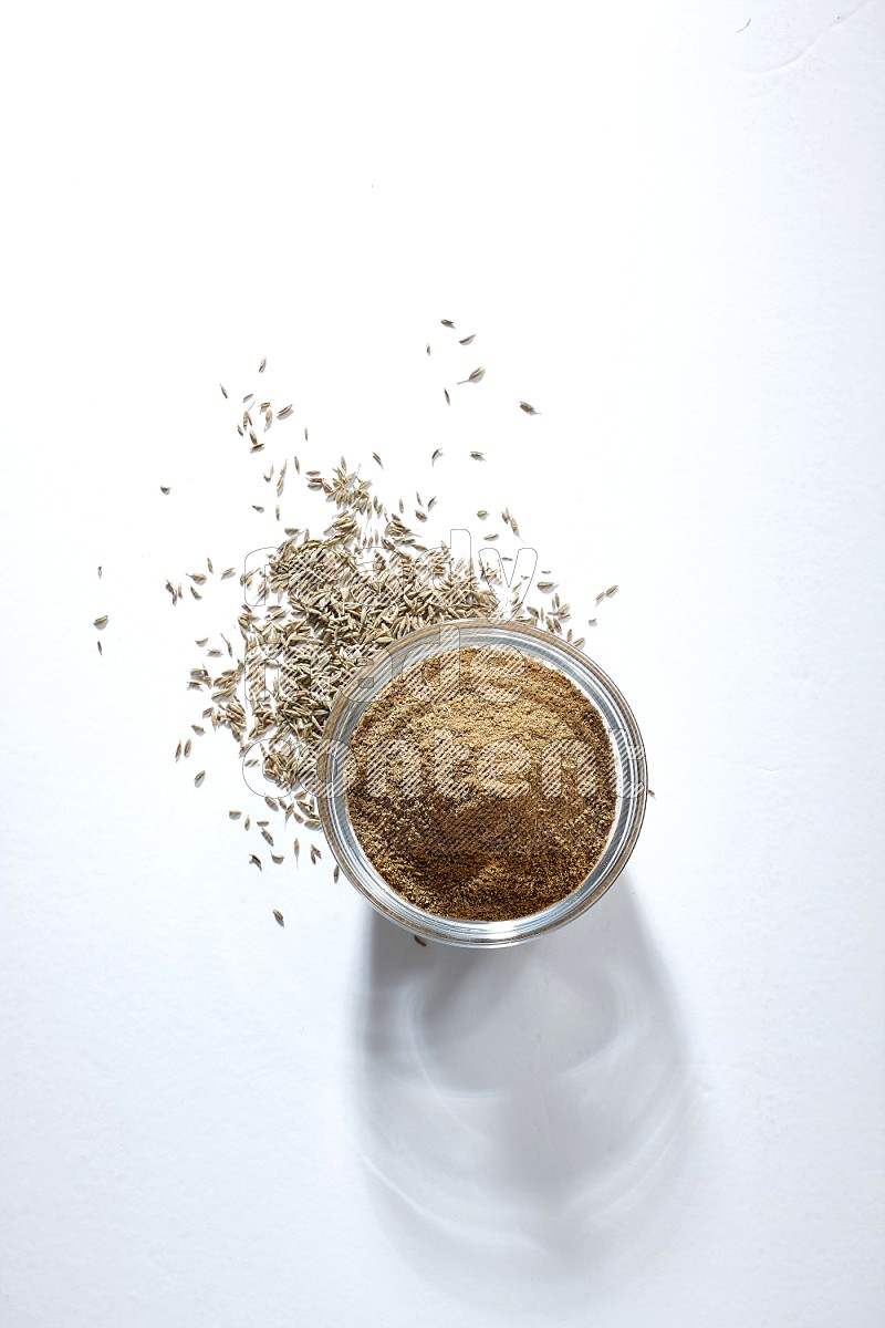 A glass bowl full of cumin powder with cumin seeds beneath it on white flooring