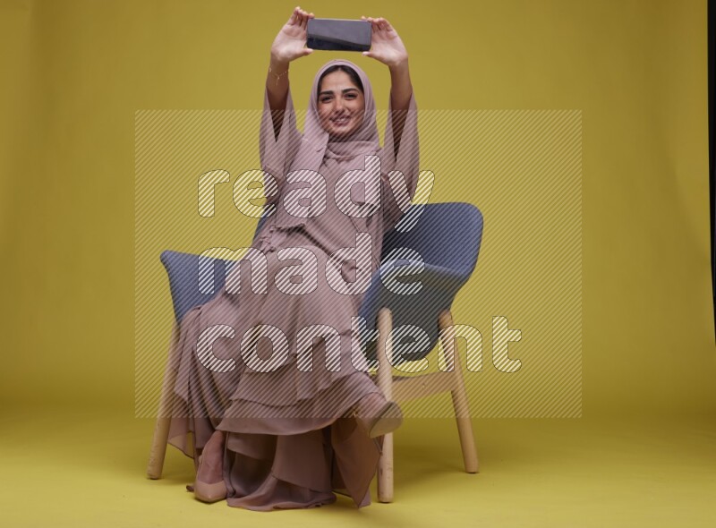 A woman Showing Her Phone Screen Setting  on a Yellow Background wearing Brown Abaya with Hijab