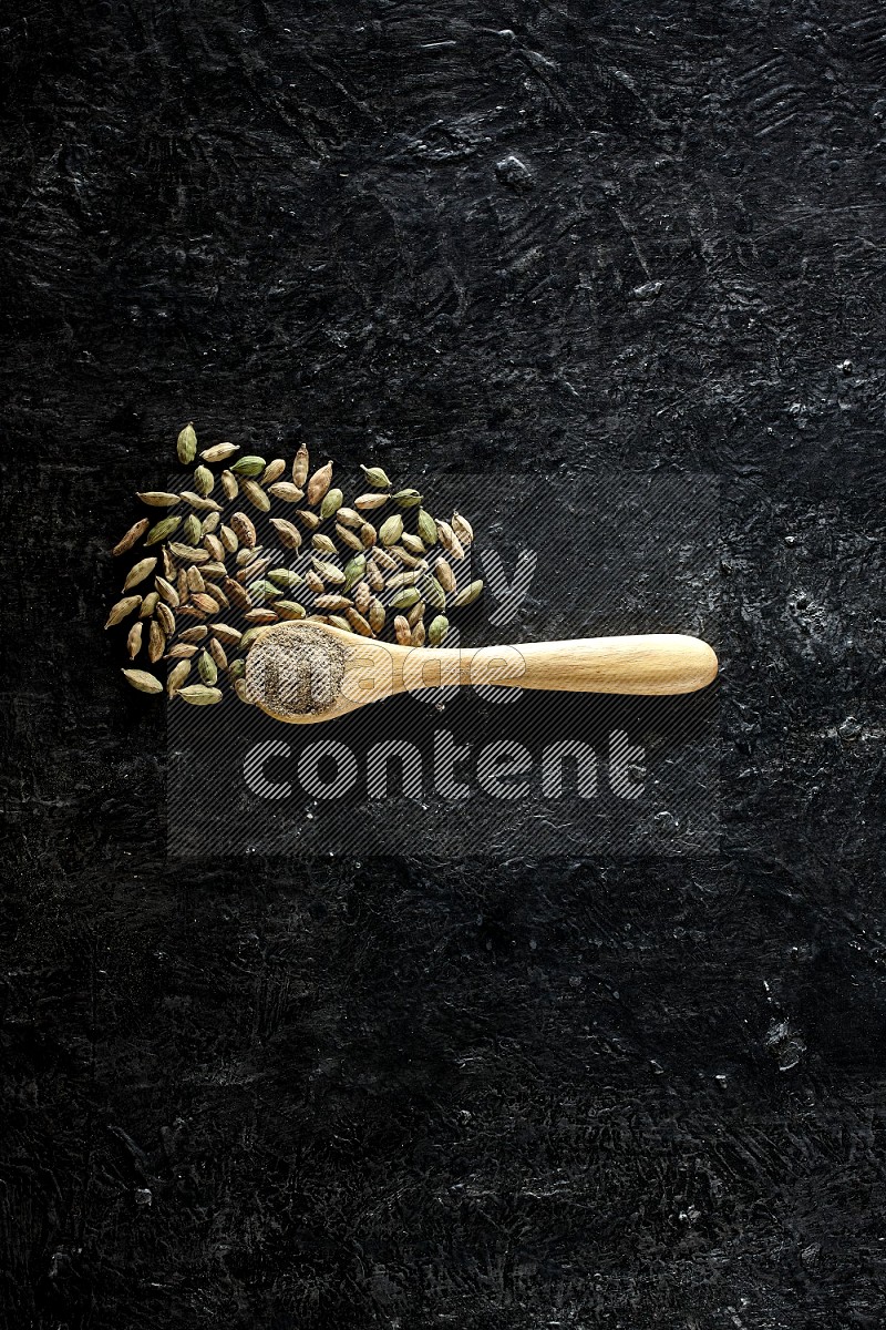 A wooden spoon full of cardamom powder and cardamom seeds next of it on textured black flooring