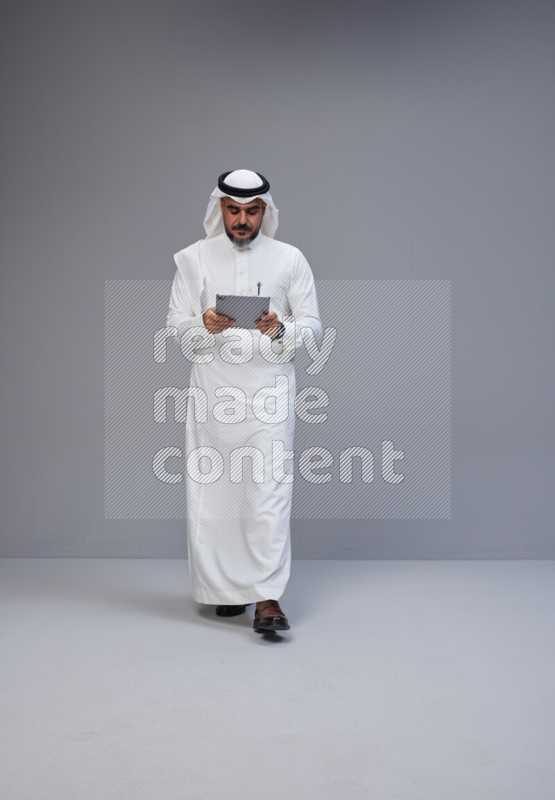 Saudi man Wearing Thob and white Shomag standing working on tablet on Gray background