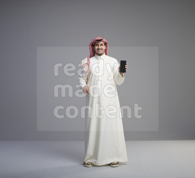 A Saudi man standing wearing thob and red shomag showing phone to camera on gray background