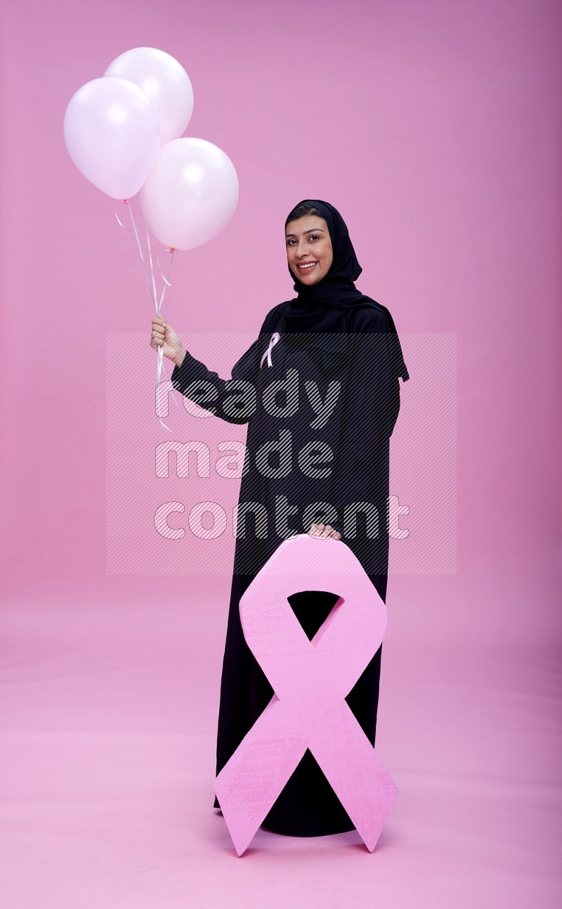Saudi woman wearing pink ribbon on Abaya standing holding balloon with awareness on plain pink background