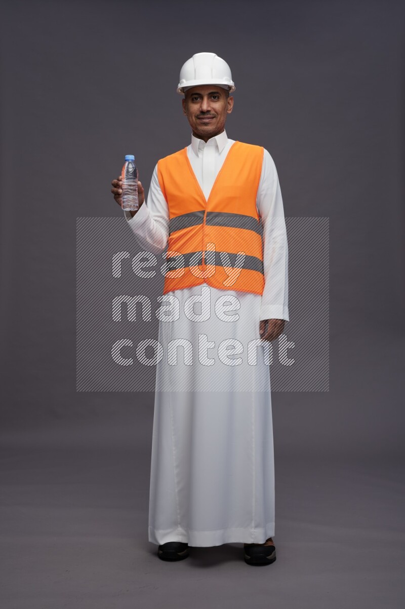Saudi man wearing thob with engineer vest standing drinking water on gray background