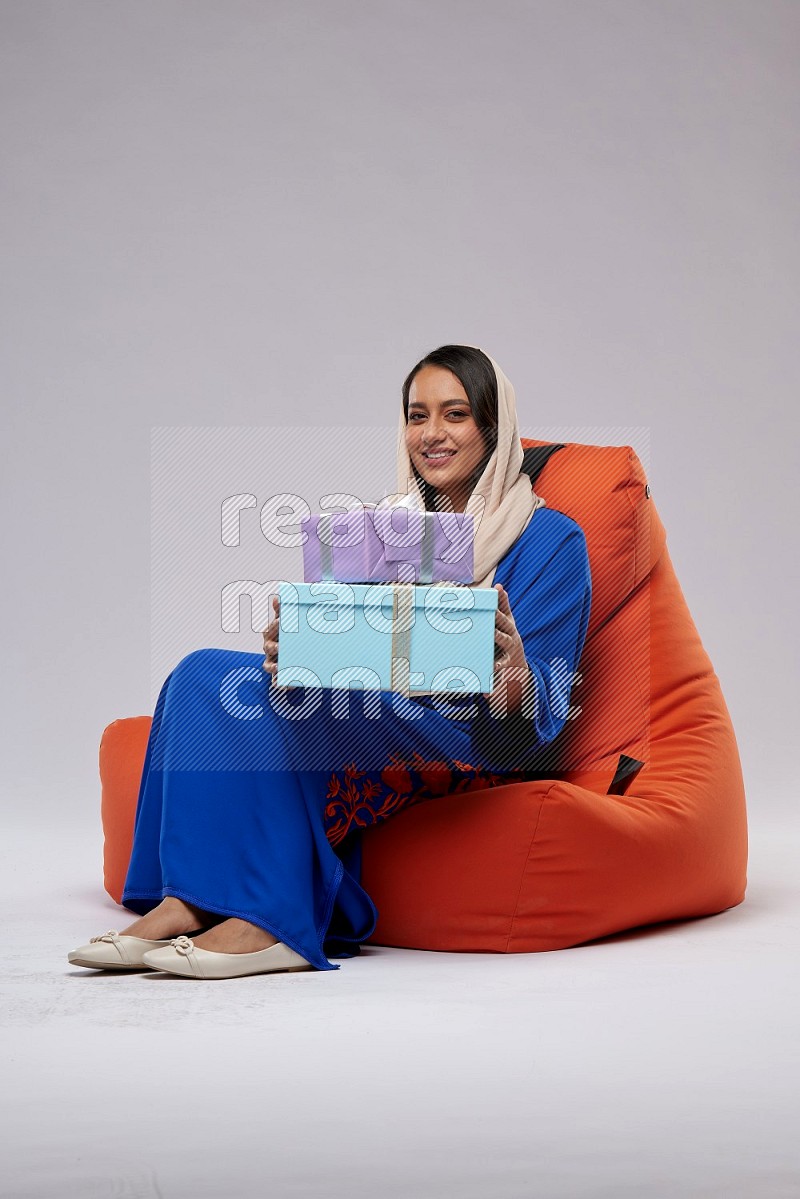 A woman sitting on an orange beanbag wearing Jalabeya holding a gift box