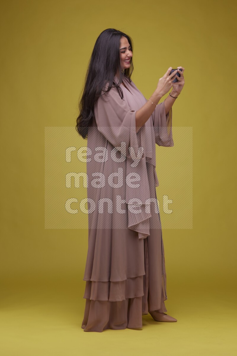 A woman Playing a Game on her smartphone on a Yellow Background wearing Brown Abaya