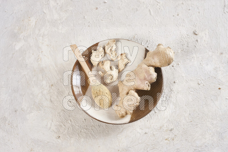 A wooden spoon full of ground ginger powder with fresh and dried ginger, all on a pottery plate on white background