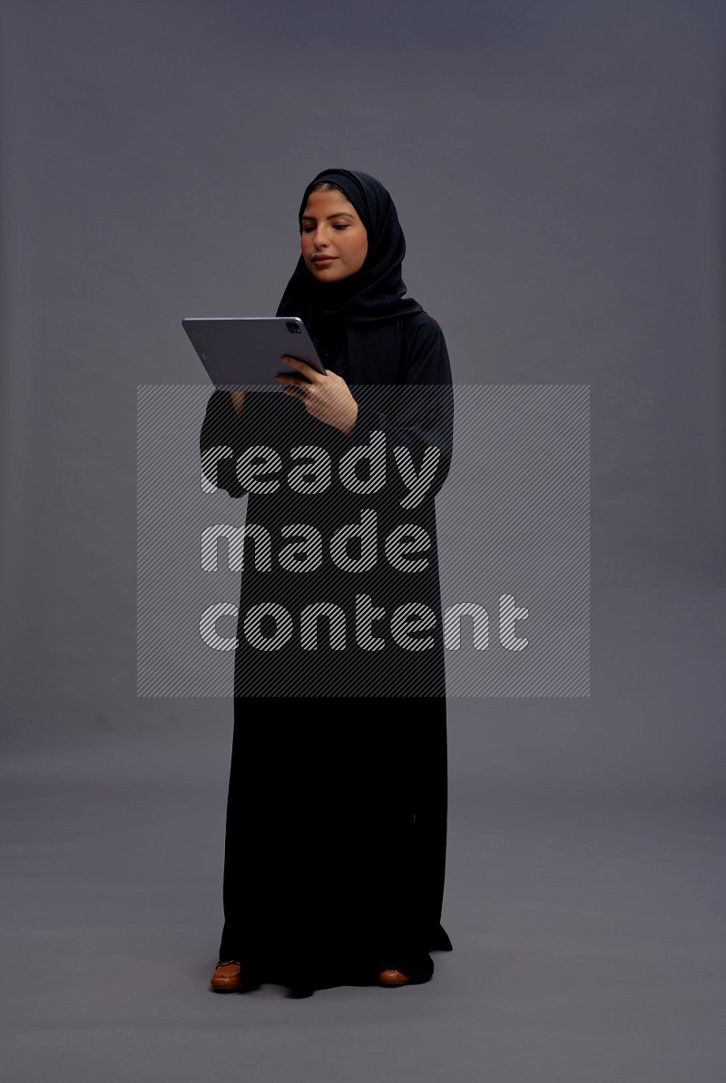 Saudi woman wearing Abaya standing working on tablet on gray background