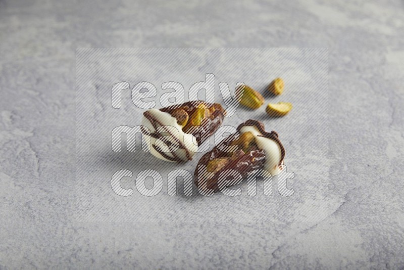 two pistachio stuffed dates covered with white chocolate with dark chocolate drizzle with unroasted pistachios on a light grey background