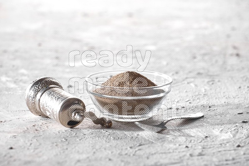 A glass bowl full of black pepper powder and a metal spoon and a turkish metal grinder on textured white flooring