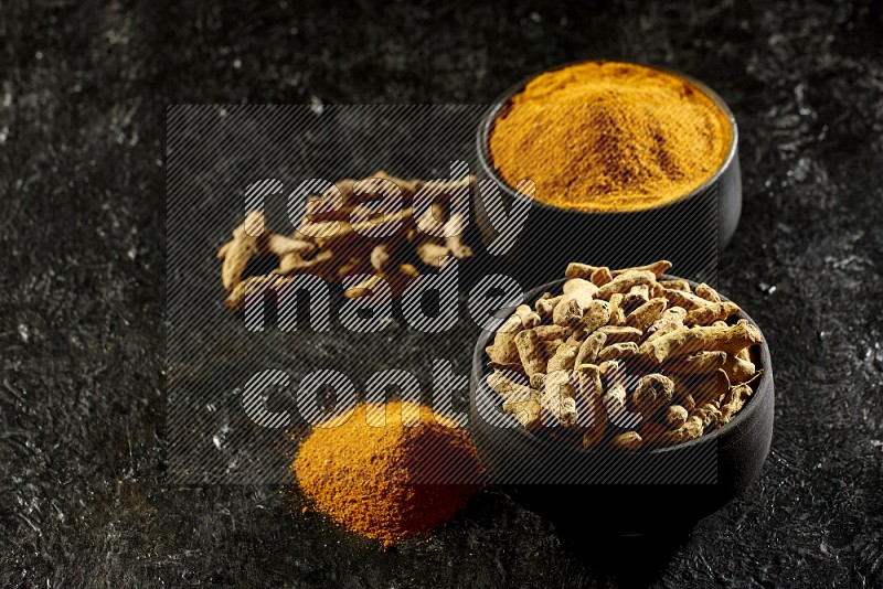 2 pottery black bowls, one full of turmeric powder and the other full of dried turmeric whole fingers on a textured black flooring