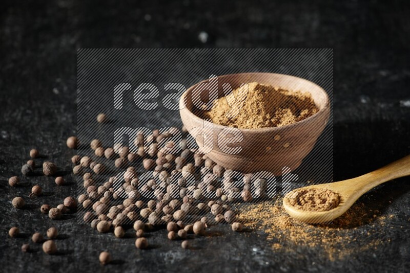 A wooden bowl and spoon full of allspice powder and whole balls spreaded on a black flooring
