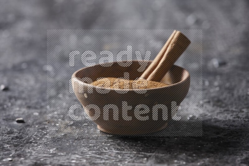 Wooden bowl full of cinnamon powder and a cinnamon stick on a textured black background