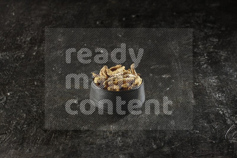 A black pottery bowl full of dried turmeric whole fingers on textured black flooring