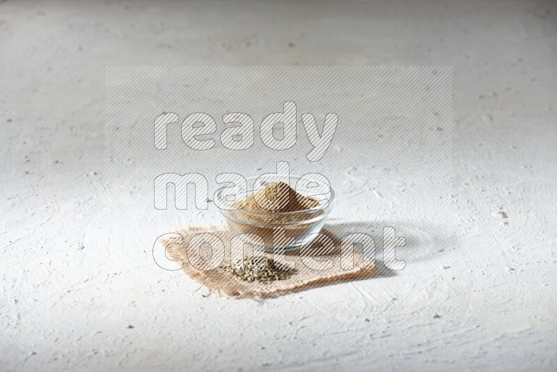 A glass bowl full of cumin powder with some of cumin seeds on burlap piece on a textured white flooring