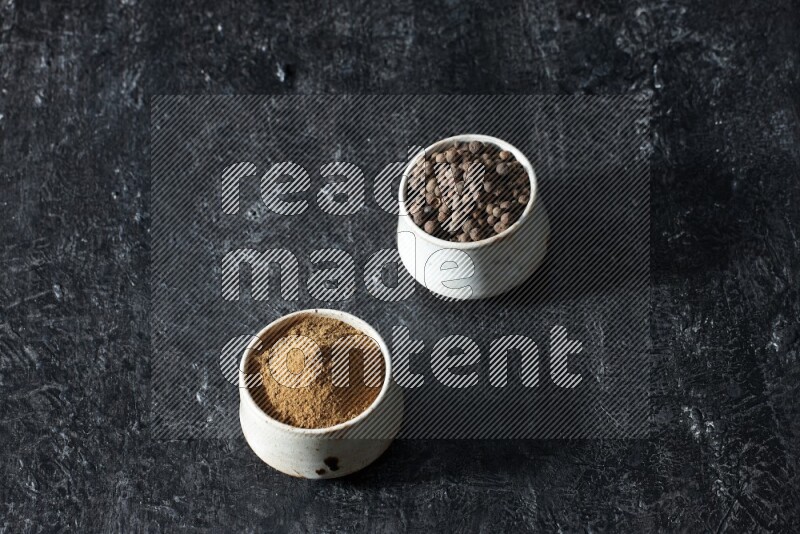 2 beige bowls, one full of allspice powder and the other full of whole balls on a textured black flooring
