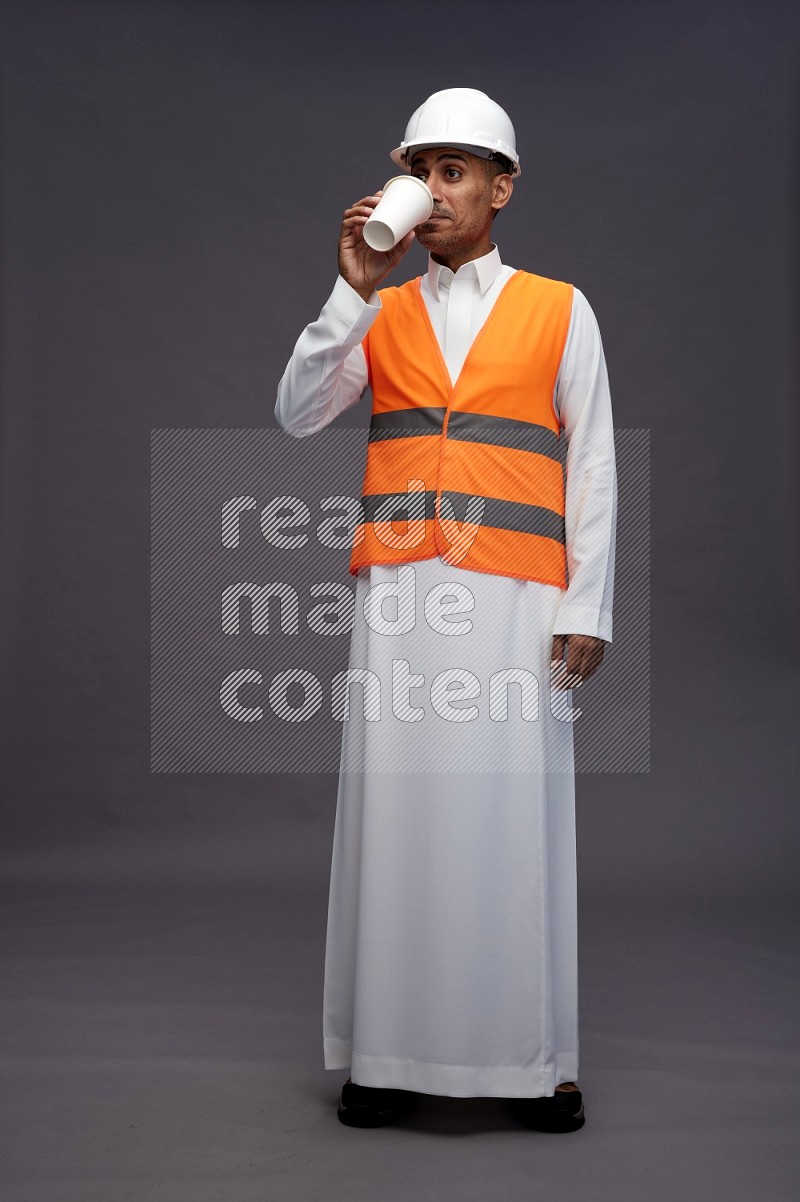 Saudi man wearing thob with engineer vest standing holding paper cup on gray background