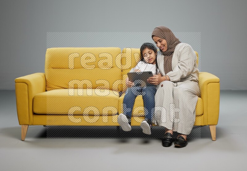 A girl with her mother on yellow sofa and watching on iPad on gray background