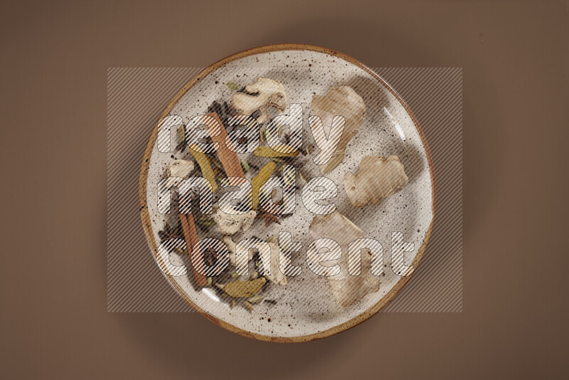 An assorted whole spices on a flat beige plate including fresh and dried ginger, cinnamon sticks, cardamom, turmeric fingers, cloves, star anise on a beige background