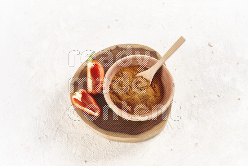 A wooden bowl full of ground paprika powder and sliced red bell pepper beside it all on a wooden tray on white background