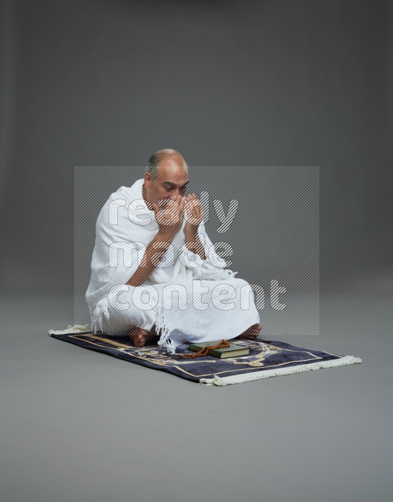 A man wearing Ehram sitting on prayer mat dua'a on gray background