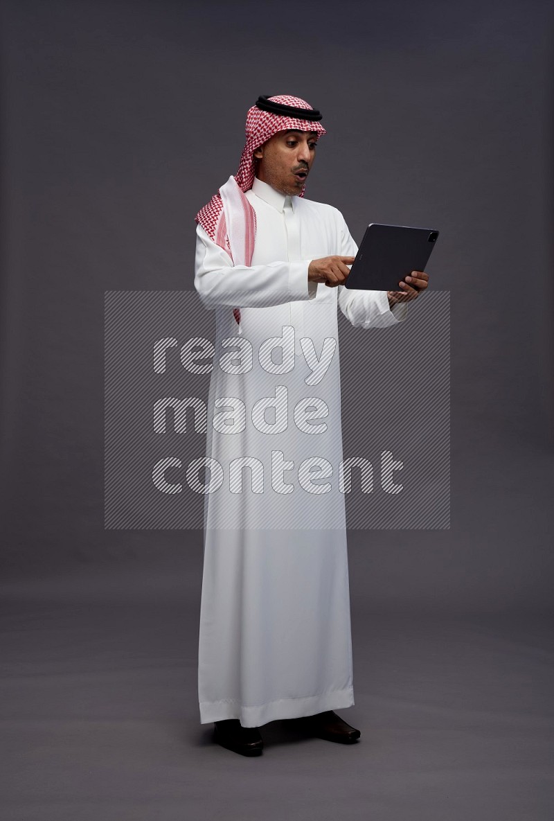 Saudi man wearing thob and shomag standing working on tablet on gray background