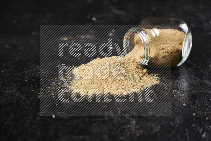 A glass jar full of ground ginger powder flipped with some spilling powder on black background