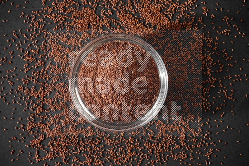 A glass bowl full of garden cress seeds surrounded by seeds on a black flooring