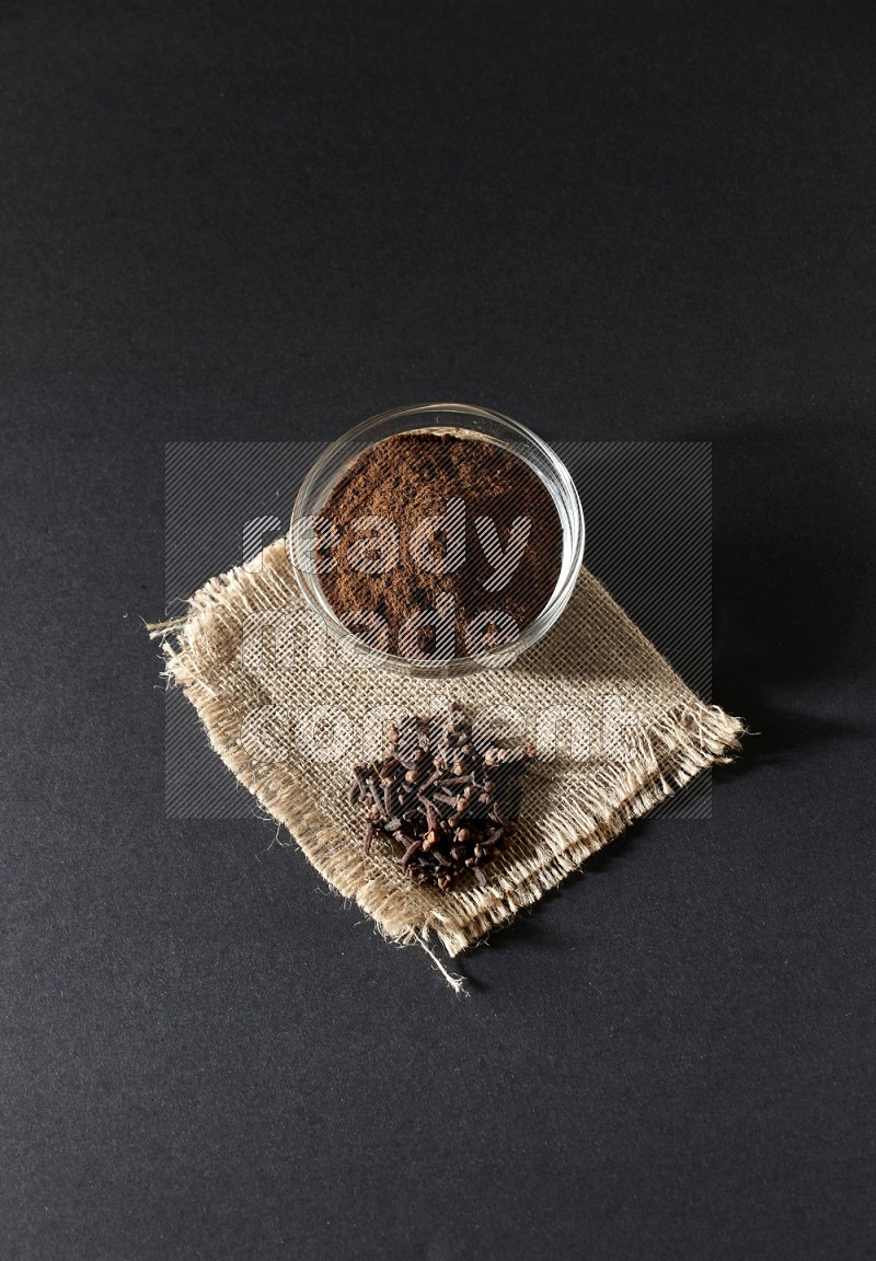 A glass bowl full of cloves powder with cloves grains on a burlap piece on a black flooring