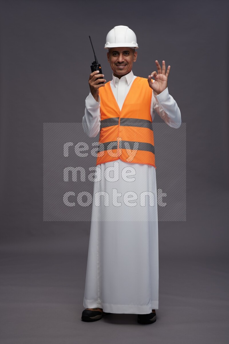 Saudi man wearing thob with engineer vest standing holding walkie-talkie on gray background
