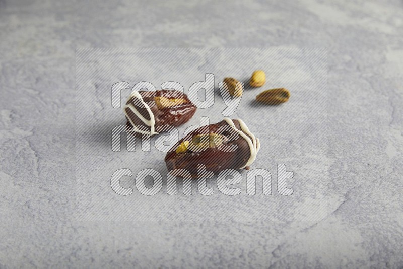 two pistachio stuffed dates covered with dark chocolate with white chocolate drizzle with unroasted pistachios on a light grey background